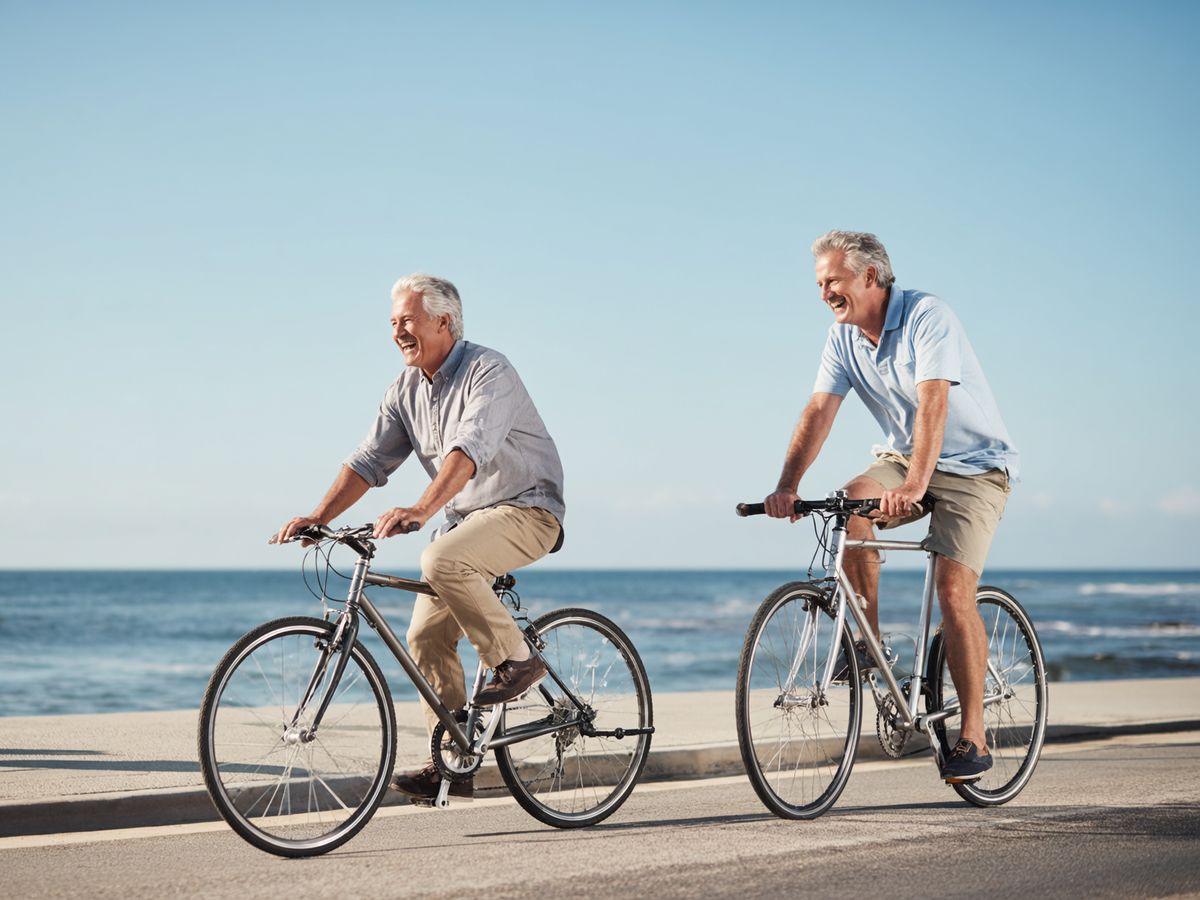 Two men in their fifties casually riding bicycles along a paved coastal path, laughing and enjoying the ocean breeze under a clear blue sky.