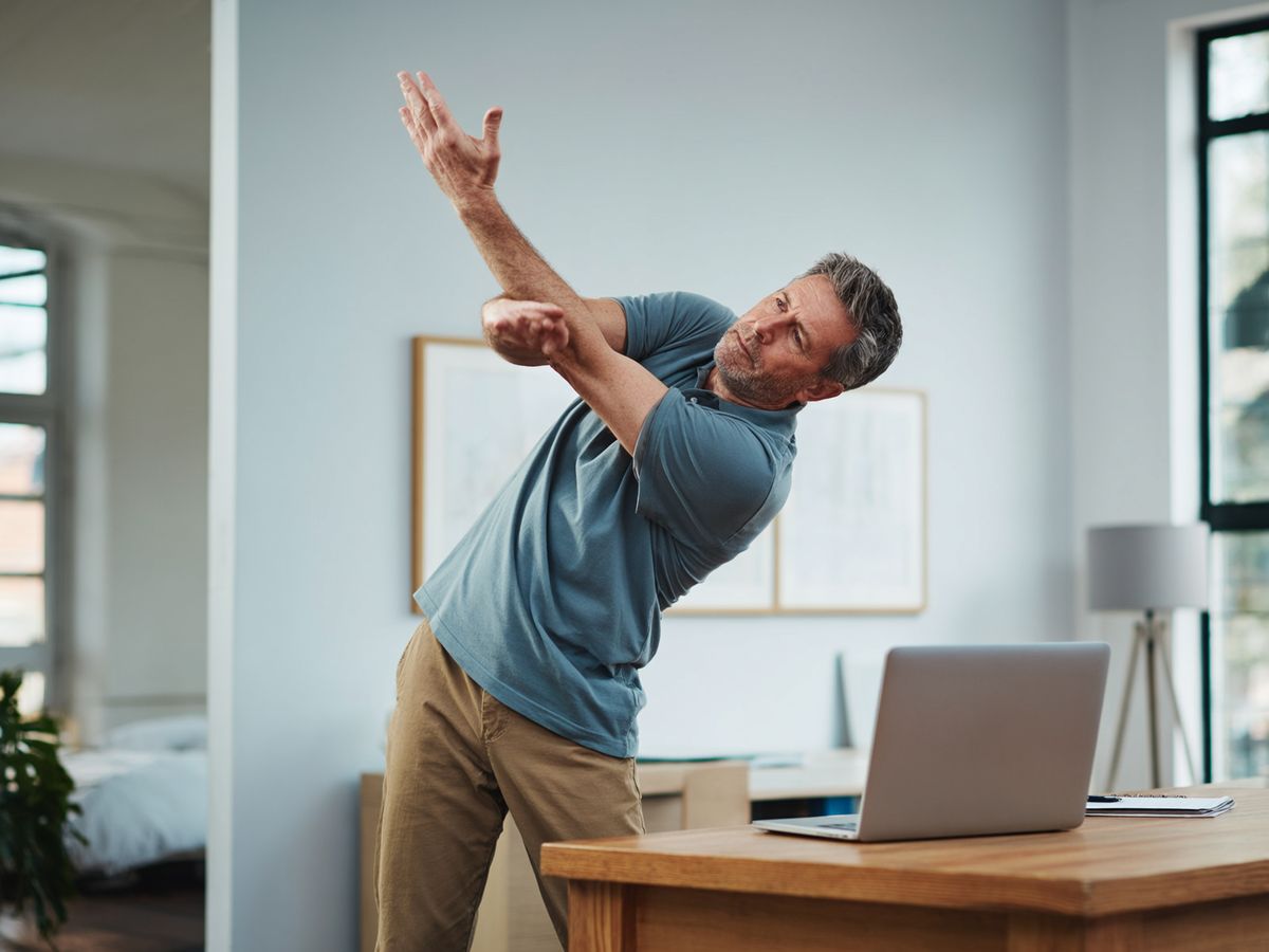 A man in his late forties in a home office, wearing casual work clothes, doing a standing torso twist stretch beside a wooden desk with a laptop.